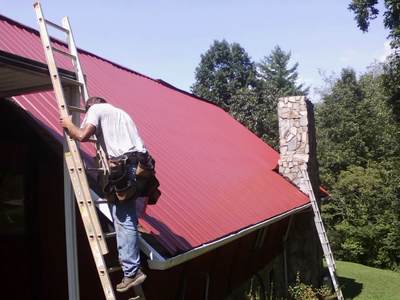 Roofer on a ladder beside a red metal roof and stone chimney — Soffit & Fascia Repair by Roofing Brothers in Amesbury
